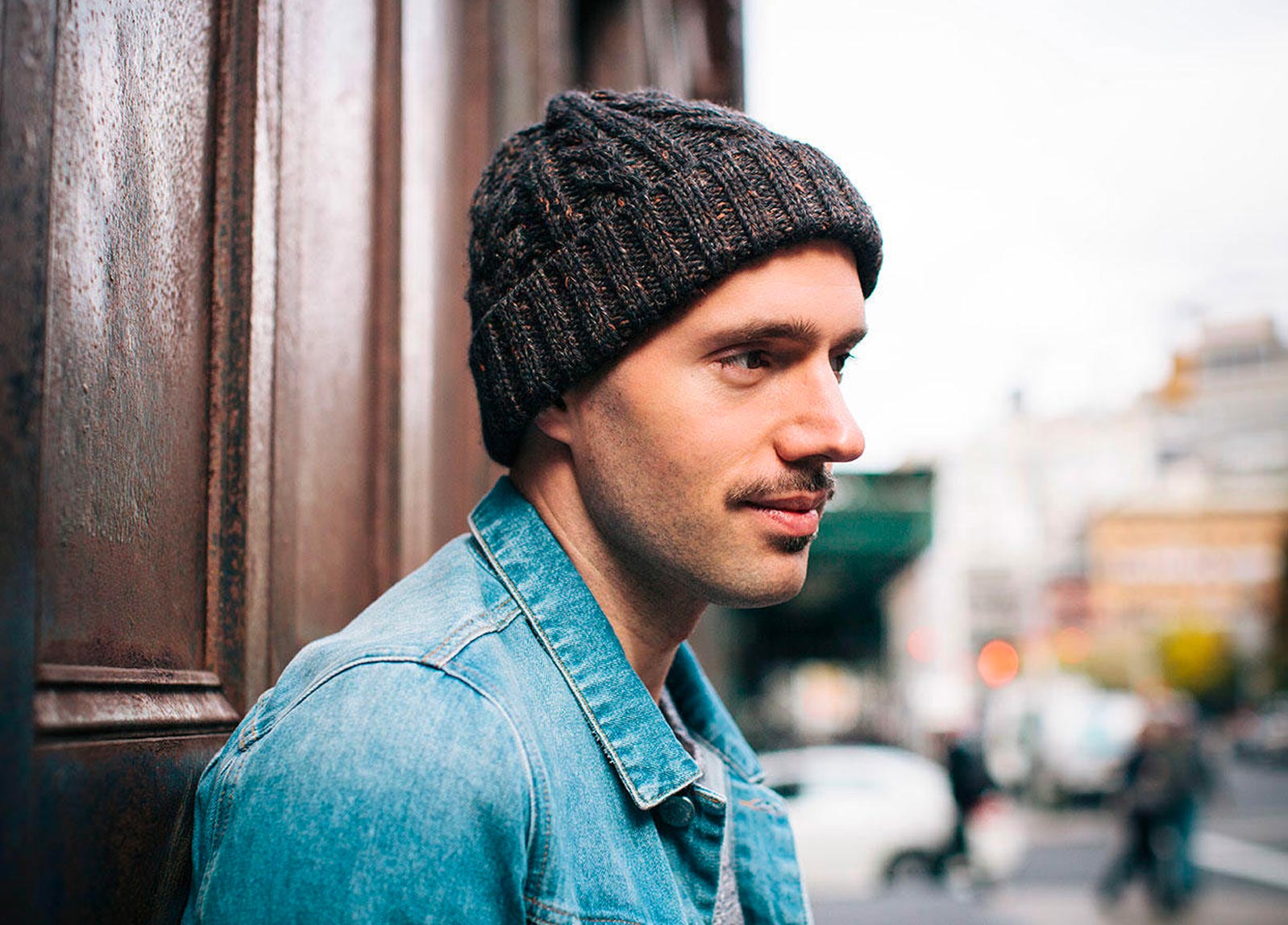 Mick Batyske wearing a jean jacket and beanie in front of a wooden door with the city in the background